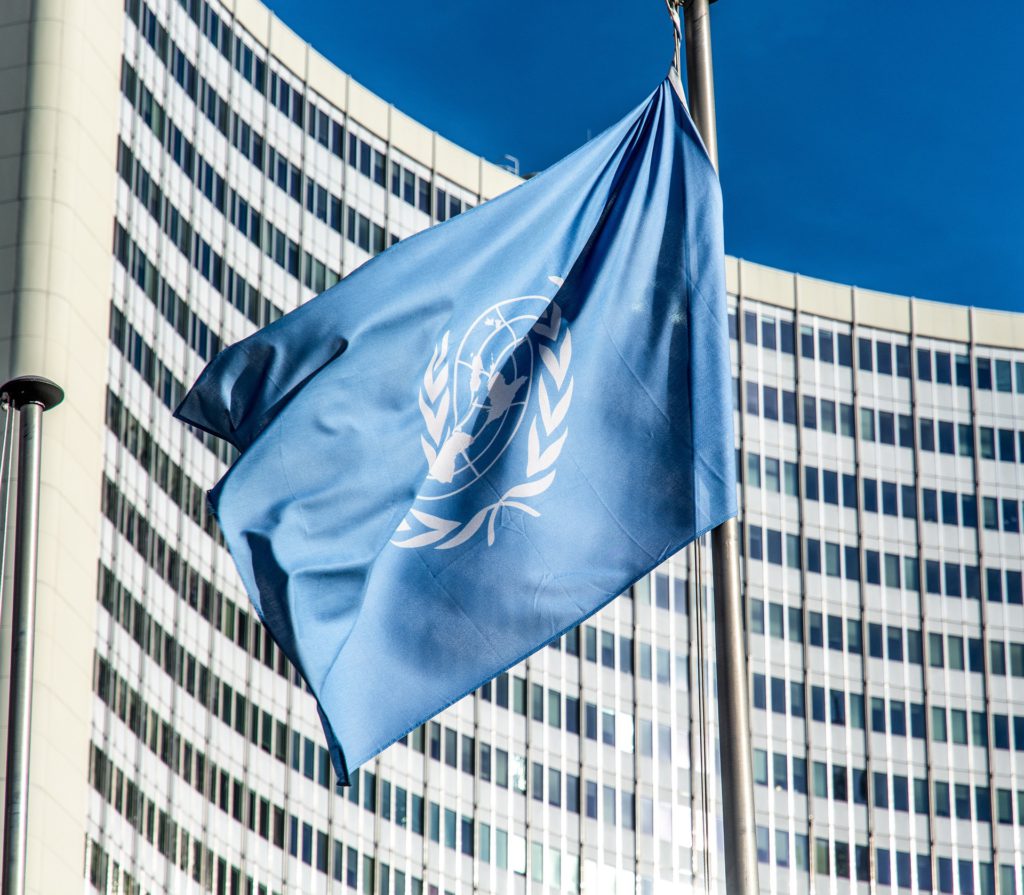 En blå flagga med FN:s symbol på. Flaggan hänger på en flaggstång framför en skyskrapa.