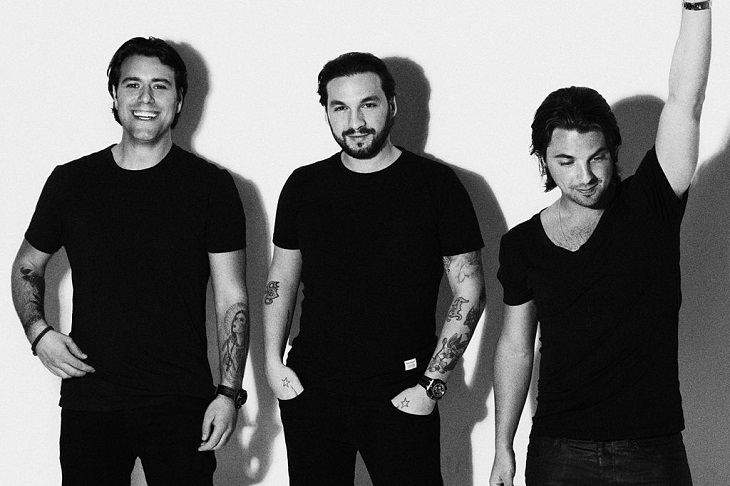 Swedish House Mafia står framför en vit vägg. De står bredvid varandra och har svarta t-shirts på sig. En skrattar, en ler lite försiktigt och en tittar bort och sträcker upp sin arm i luften.