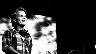 Avicii står bakom ett mixerbord. Han har en rutig skjorta och halvlångt hår som är struket bakåt. Han tittar ut mot publiken och ler.