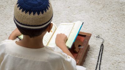 Ett barn sitter med ryggen mot oss. Framför sig har det en uppslagen koran på en bricka. Bredvid ligger ett ban med pärlor. Barnet har en virkad lite mössa på sig.