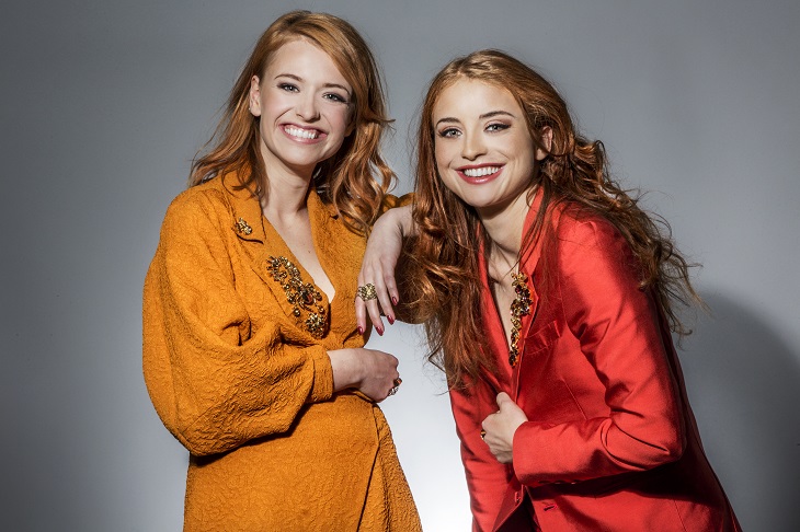 Clara och Henry ler stort mot kameran. Båda har långt svallaande hår och mycket smink. De håller i sina jackor så de ska vara stängda. De har stora glimrande stenar på sina jackor och många ringar.
